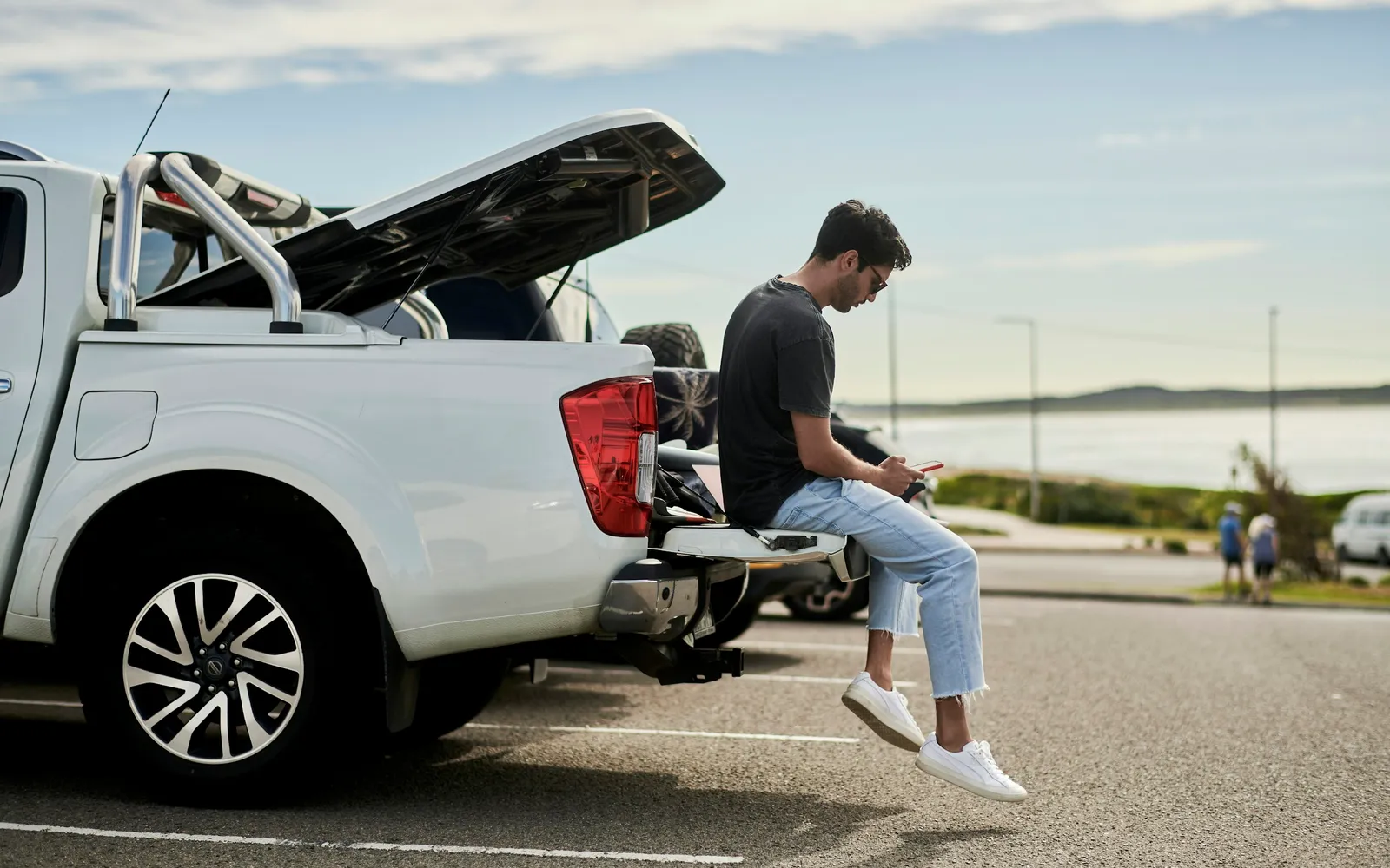 A man sits on the tailgate of a ute in a car park by the sea, looking at his phone.