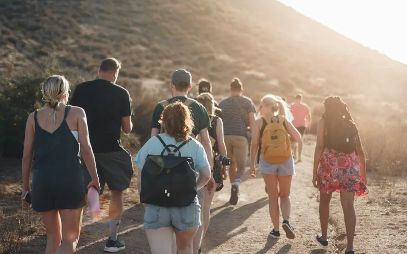 A group with daypacks walks a dirt trail into the low evening sun, seen from behind.