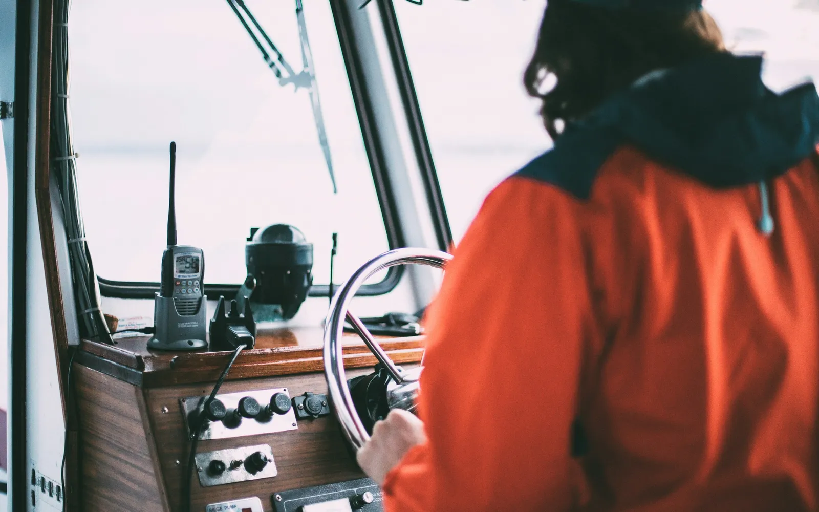 A skipper in an orange jacket steers a small boat from the wheelhouse.