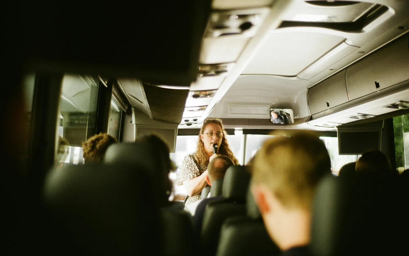 A guide talks into a microphone at the front of a coach while passengers listen from their seats.