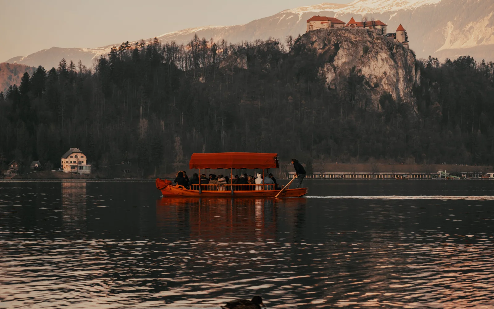 A guide stands at the stern of a canopied boat full of guests and rows them across a lake below a castle in the late light.