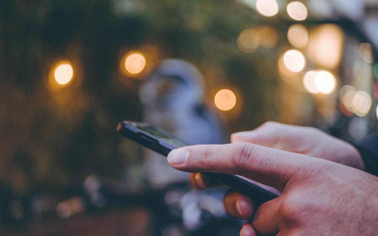 A hand holds a phone at dusk, one finger about to tap the screen, with warm lights blurred behind it.