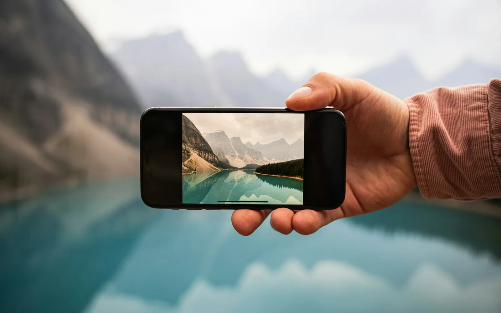 A hand holds up a phone in front of a turquoise mountain lake, the same view sharp on its screen.