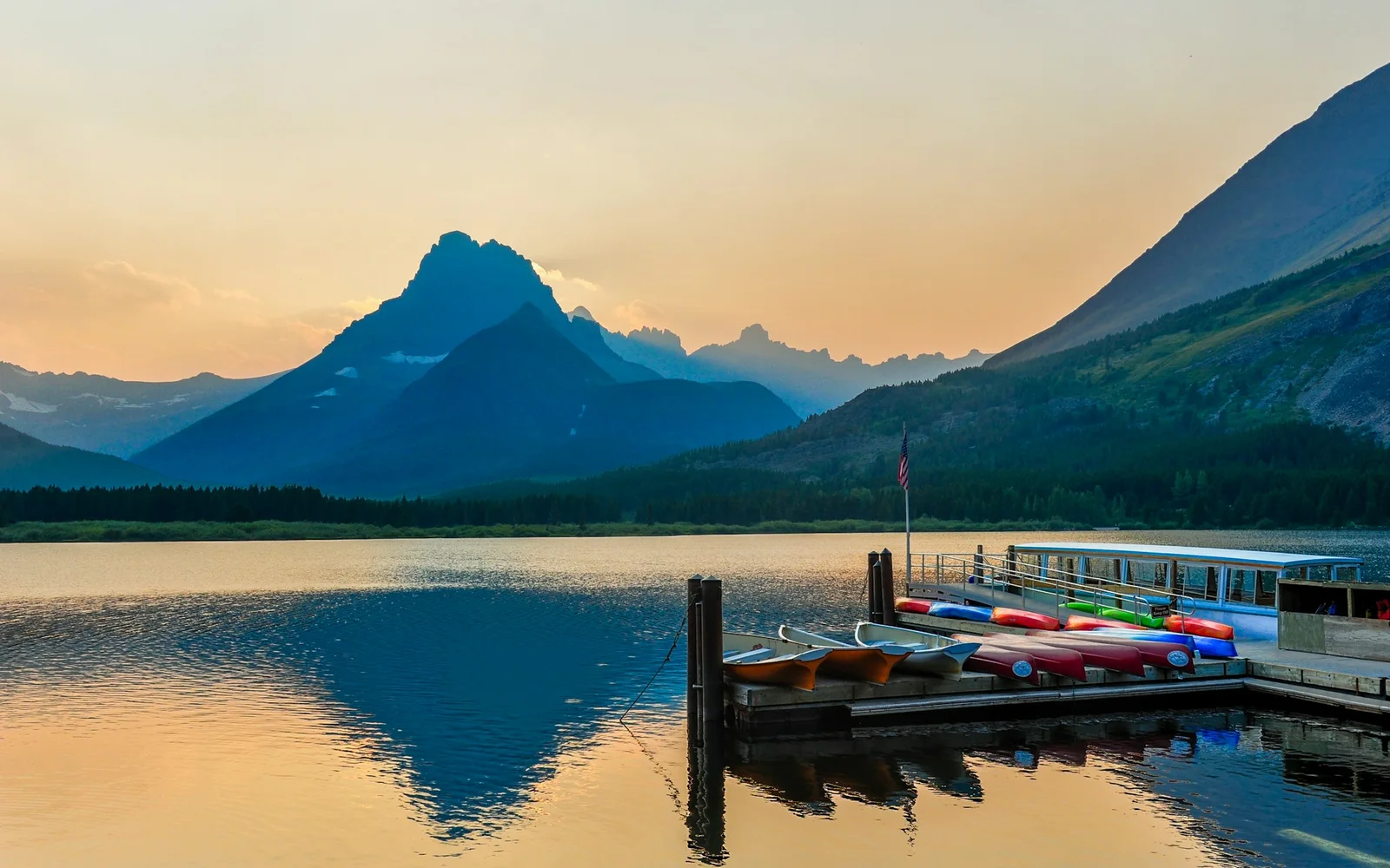 An empty tour boat waits at a lake dock at dusk beside a row of canoes and kayaks, with mountains behind.